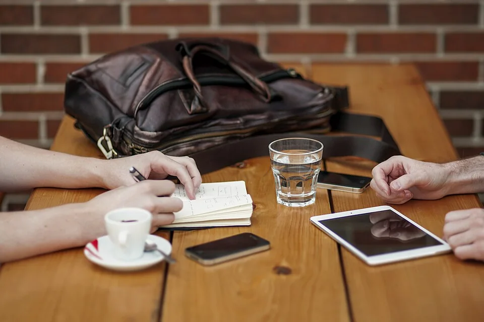 Two people’s hands beside a notebook, smartphone, tablet and drinks on a table.