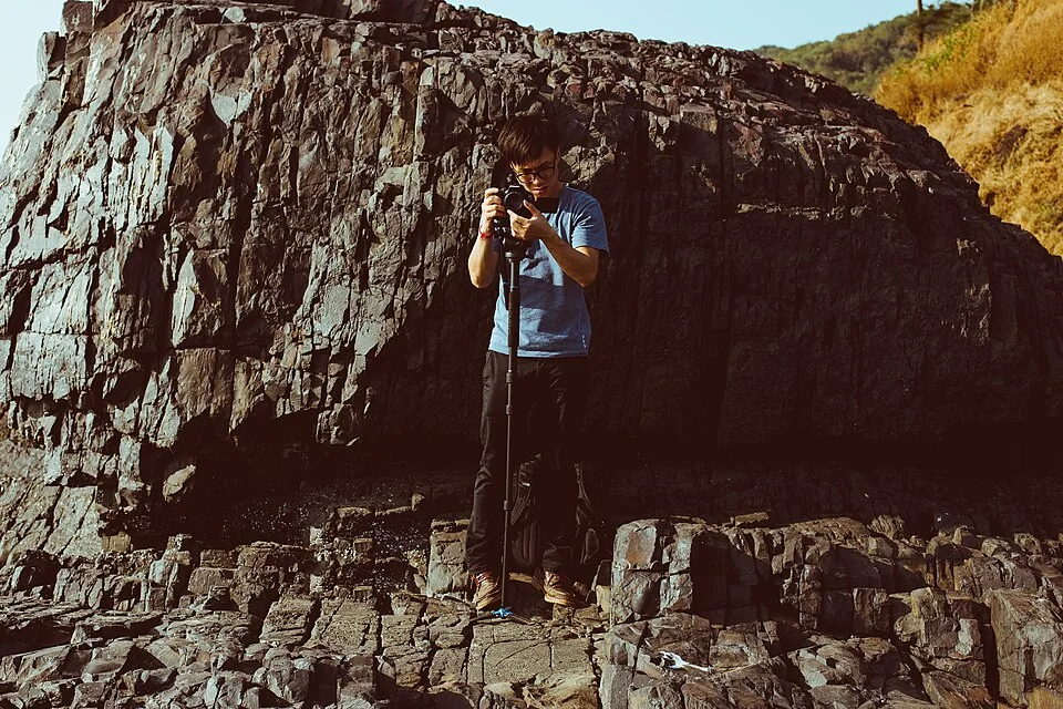A young man films with a camera on a monopod in front of a dark rock face.