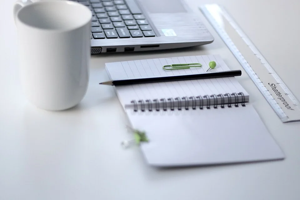 A ruler, notebook and pen beside a laptop on a bright desk.
